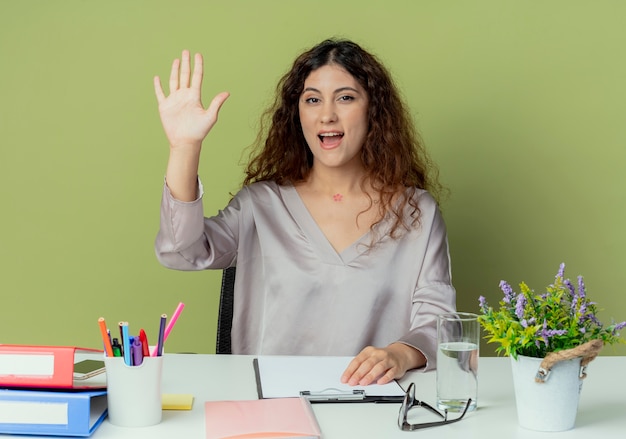 Lavoratore di ufficio femminile grazioso giovane allegro che si siede allo scrittorio con gli strumenti dell'ufficio che mostrano il gesto di ciao isolato su fondo verde oliva