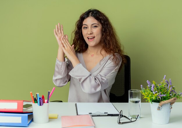 Lavoratore di ufficio femminile grazioso giovane allegro che si siede allo scrittorio con la mano d'applauso degli strumenti dell'ufficio isolata su fondo verde oliva