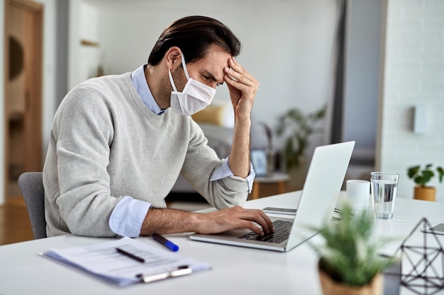 Lavoratore freelance preoccupato che indossa una maschera protettiva mentre legge e-mail problematiche su un computer e lavora a casa durante l'epidemia di virus