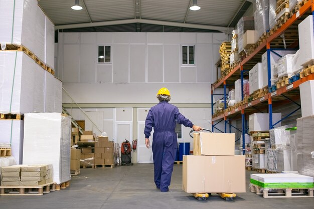 Lavoratore logistico senior in elmetto protettivo e uniforme a piedi in magazzino, presa della tavolozza con ruote Vista posteriore, a figura intera. Concetto di lavoro e logistica