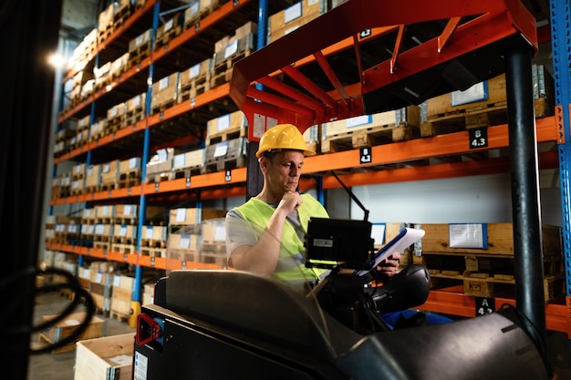 Lavoratore manuale seduto in un carrello elevatore e leggere documenti mentre lavora in magazzino