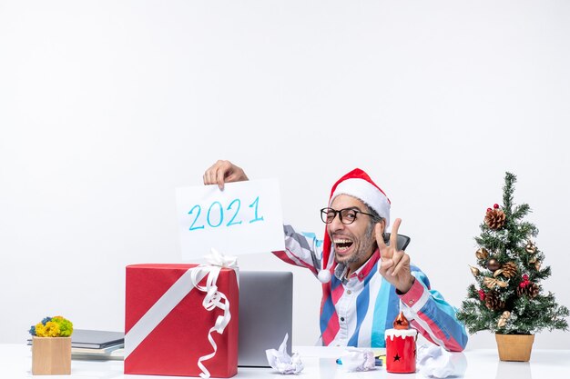 Lavoratore maschio di vista frontale seduto nel suo posto di lavoro che tiene felicemente un foglio di carta con il numero 2021, concetto del nuovo anno