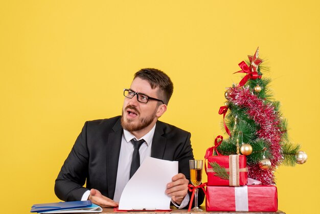 Lavoratore maschio vista frontale dietro il suo tavolo con regali e albero di Natale su giallo