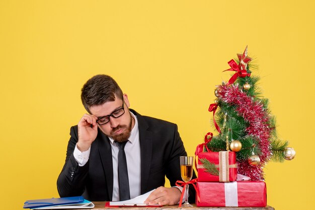 Lavoratore maschio vista frontale dietro il suo tavolo con regali e albero di Natale su giallo