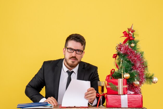 Lavoratore maschio vista frontale dietro il suo tavolo con regali e albero di Natale su giallo