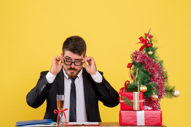 Lavoratore maschio vista frontale dietro il suo tavolo con regali e albero di Natale su giallo