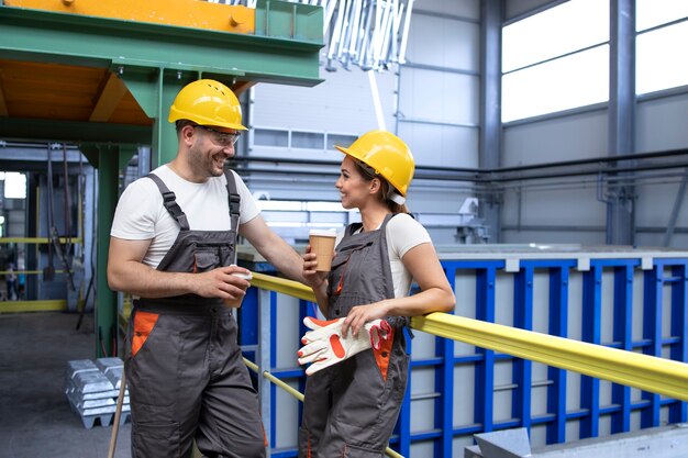 Lavoratori dell'industria in uniforme e attrezzature di sicurezza che si rilassano in una pausa bevendo caffè e parlando all'interno della fabbrica