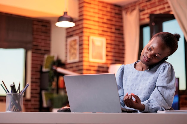 Lavoratrice remota femminile che risponde alla telefonata e lavora al computer portatile nel moderno ufficio a casa. Donna che discute di piani aziendali con i colleghi dell'azienda su smartphone, freelance che chatta con i clienti