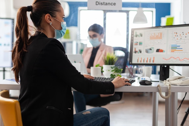 Lavoro di squadra che indossa una maschera medica per prevenire l'infezione da coronavirus che lavora alla strategia di gestione nell'ufficio aziendale. Donne d'affari che pianificano la presentazione dell'azienda. Grafico finanziario sullo schermo