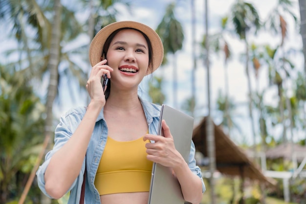 Lavoro e vacanza asiatico femmina nomade digitale stile di vita millenario tenere in mano smartphone conversazione con all'estero cliente distanza incontro asiatico femminile panno casual camminare parlando sulla spiaggia oceano