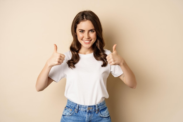 Lavoro eccellente. Felice giovane donna che mostra i pollici in su in segno di approvazione e sorridente, in piedi in t-shrit su sfondo beige.