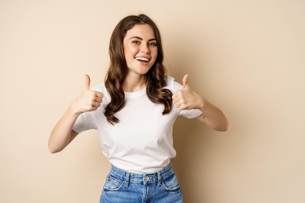 Lavoro eccellente. Felice giovane donna che mostra i pollici in su in segno di approvazione e sorridente, in piedi in t-shrit su sfondo beige.