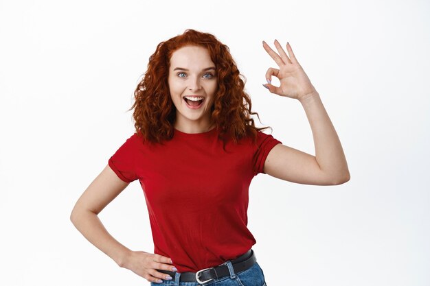 Lavoro eccellente. La ragazza rossa contenta con i capelli ricci dice bene sì, mostrando il segno ok e sorridendo soddisfatto, approva la buona cosa, in piedi sul muro bianco