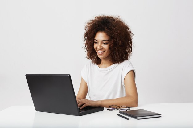 Lavoro sorridente della donna di affari africana al computer portatile sopra la parete bianca.