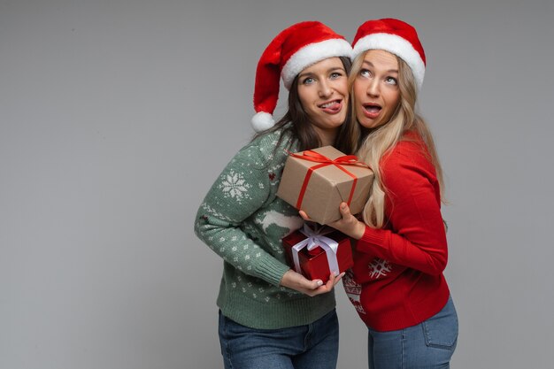 Le amiche attraenti con cappelli natalizi rossi e bianchi tengono un regalo l'una per l'altra