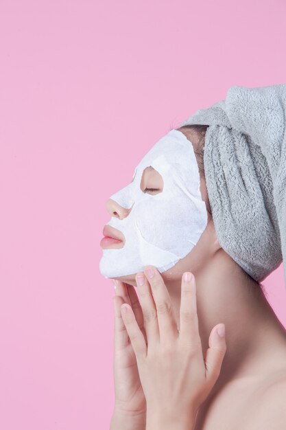 Le belle donne asiatiche stanno usando il fronte della maschera facciale sullo strato su un fondo rosa.