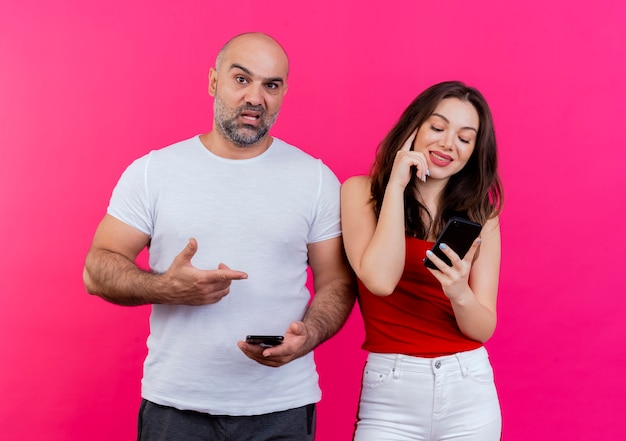 Le coppie adulte che tengono entrambi i telefoni cellulari hanno impressionato l'uomo che guarda indicando la donna e la donna felice che tocca la testa che guarda il telefono ha impressionato l'uomo