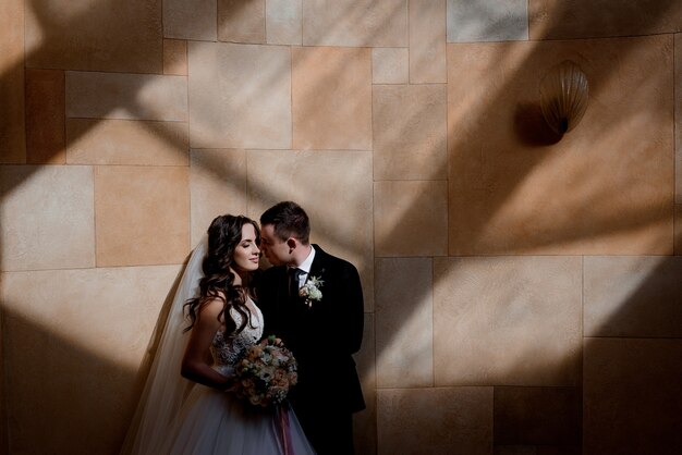 Le coppie di nozze stanno stando vicino alla parete nei raggi del sole e stanno quasi baciando, concetto del matrimonio