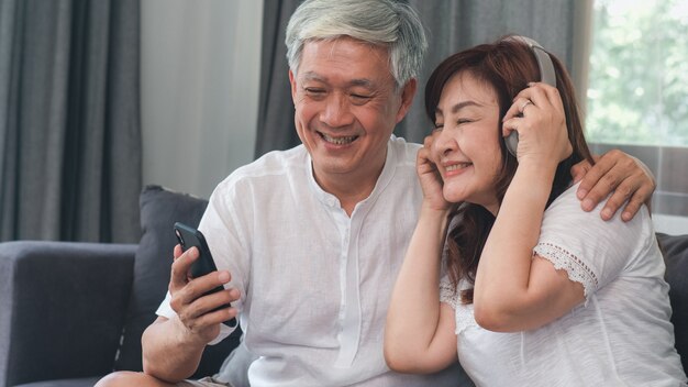 Le coppie senior asiatiche si rilassano a casa. La cuffia felice senior asiatica asiatica di usura dei nonni, del marito e della moglie facendo uso del telefono cellulare ascolta musica mentre si trova sul sofà nel concetto del salone a casa.