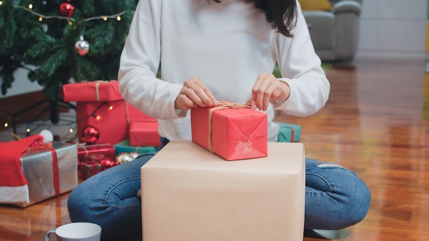 Le donne asiatiche celebrano il festival di Natale. Il maglione teenager femminile di usura e il cappello di Natale si rilassano i regali avvolgenti felici vicino all'albero di Natale godono insieme delle vacanze invernali di natale in salone a casa.