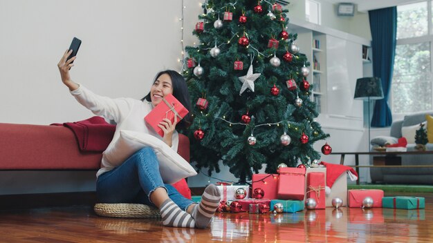 Le donne asiatiche celebrano il festival di Natale. L'adolescente femminile si rilassa il regalo felice della tenuta e l'utilizzo del selfie dello smartphone con l'albero di Natale gode delle vacanze invernali di natale nel salone a casa.