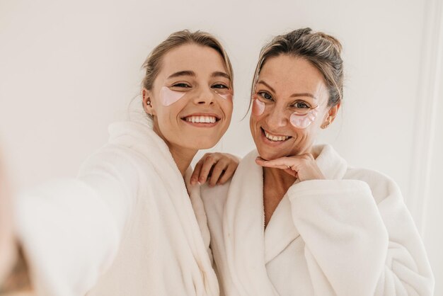 Le donne caucasiche felici usano bende per gli occhi, indossano accappatoi e guardano la fotocamera su sfondo bianco. Concetto di preservare la giovinezza