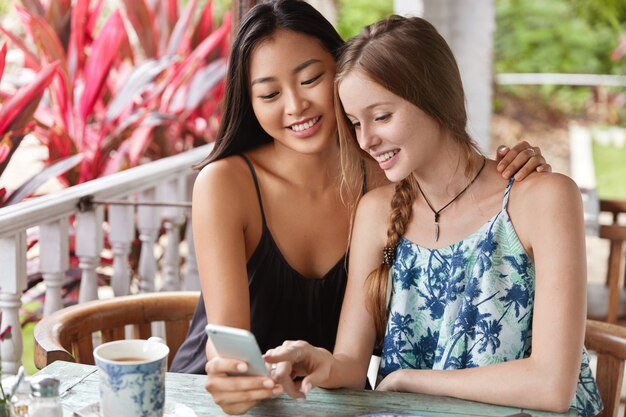 Le donne multietniche leggono la notifica con notizie positive sul cellulare, trascorrono il tempo libero in un bar con terrazza, si abbracciano, hanno espressioni felici.