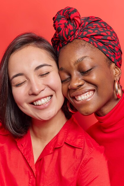 le donne stanno vicine l'una all'altra tengono gli occhi chiusi sorridono con i denti vestiti con abiti casual si divertono a trascorrere il tempo libero insieme isolate sul rosso. Concetto di emozioni positive