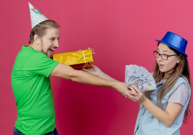 Le giovani coppie allegre che portano i cappelli del partito si guardano l'un l'altro che tiene il contenitore di regalo ed i soldi isolati sulla parete rosa