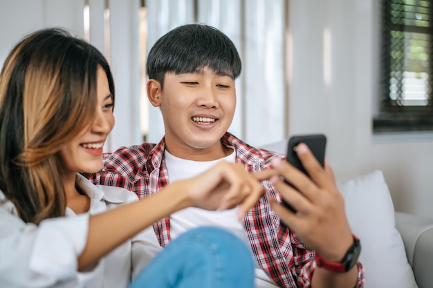 Le giovani coppie che si siedono sul divano in soggiorno e usano lo smartphone per giocare insieme parlano e ridono con felicità concetto di famiglia felicex9