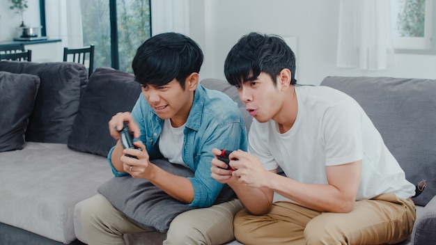 Le giovani coppie gay asiatiche giocano a casa, uomini teenager coreani LGBTQ che utilizzano la leva di comando che ha momento felice divertente insieme sul sofà nel salone a casa.
