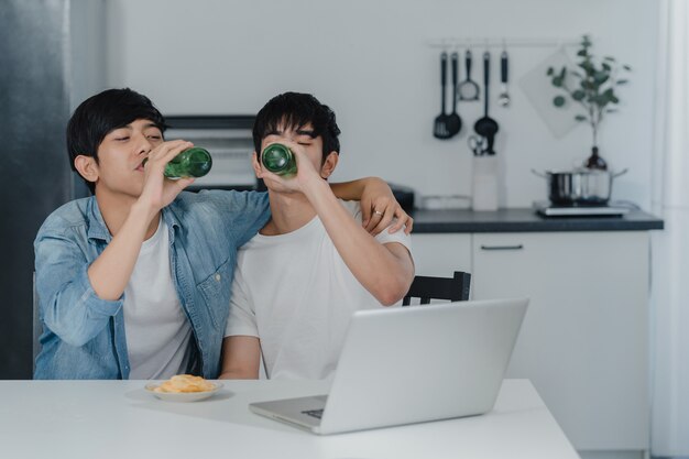 Le giovani coppie gay bevono la birra mentre utilizzano il computer portatile del computer a casa moderna. Gli uomini asiatici di LGBTQ si rilassano felici usando la tecnologia gioca insieme ai social media mentre sono seduti a tavola in cucina a casa.