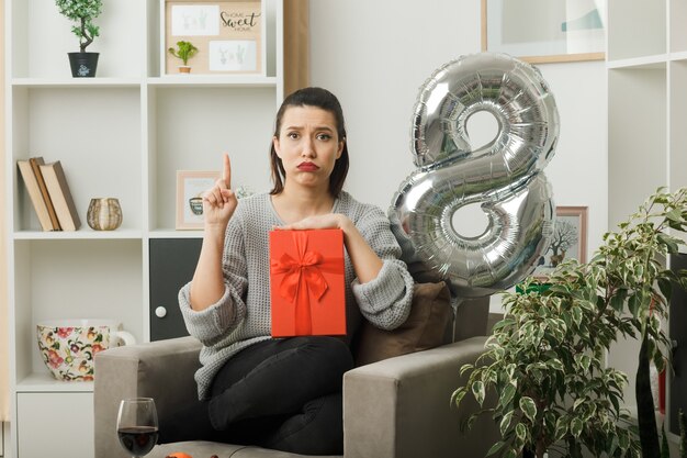 Le guance gonfie e tristi puntano su una bella ragazza durante la felice giornata delle donne con in mano un regalo seduto sulla poltrona in soggiorno