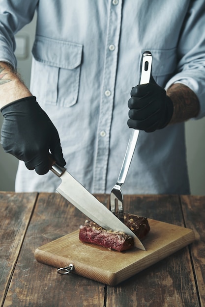 Le mani del macellaio tatuato in guanti neri tengono la fetta tagliata del coltello di carne alla griglia sulla tavola di legno