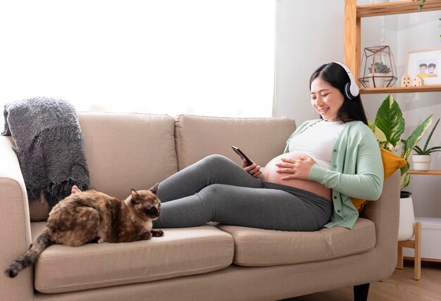 Le mani della bella donna incinta asiatica accarezzano la pancia e ascoltano musica con le cuffie che si siedono sul divano a casa Preparazione alla gravidanza e concetto di aspettativa