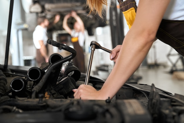 Le mani della donna che riparano automobile sotto il cappuccio con lo strumento.