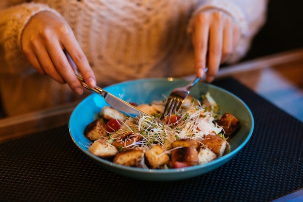 Le mani delle donne con forchetta e coltello per insalata caesar servita in una ciotola.