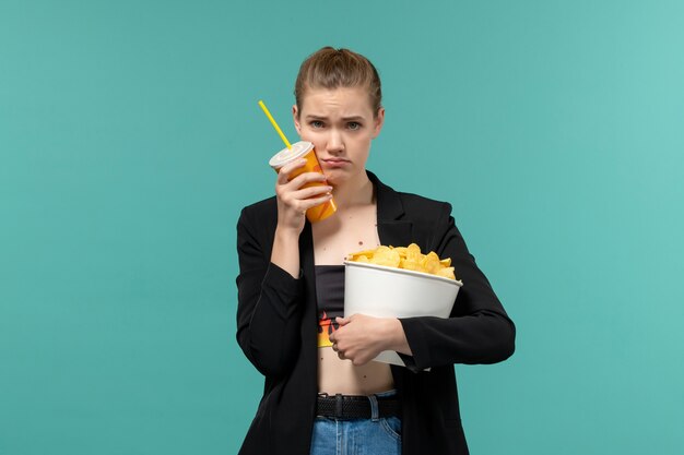 Le patatine fritte della tenuta della giovane femmina di vista frontale bevono e guardano il film sulla superficie blu-chiaro