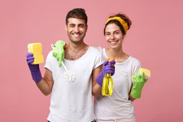 Le pulizie, i doveri domestici e il concetto di lavoro di squadra. Bella giovane famiglia europea che condivide le faccende domestiche: donna con spugna e scopino per pulire il bagno mentre l'uomo lava i vetri con lo spray