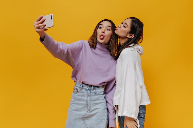 Le ragazze emotive felici si divertono e dimostrano le lingue Le giovani donne asiatiche si fanno selfie e fanno facce buffe su sfondo arancione