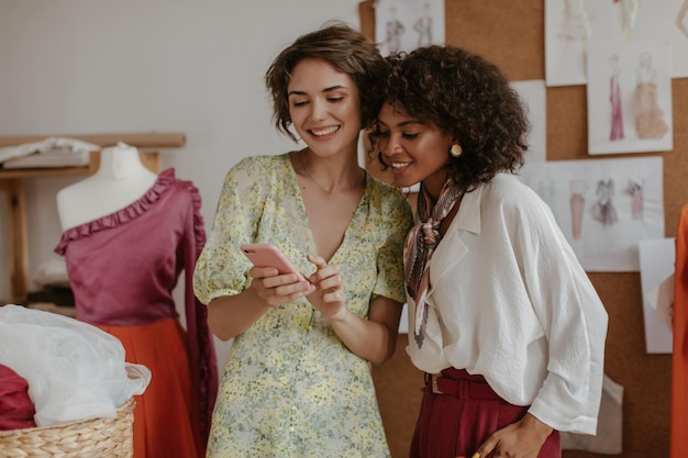 Le ragazze graziose guardano lo schermo del telefono Le donne in eleganti abiti estivi sorridono e posano nell'accogliente ufficio dello stilista