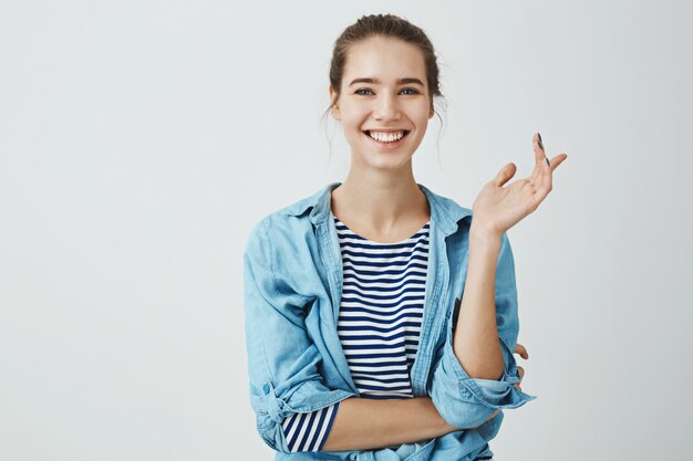 Le risate risolvono i problemi quotidiani. Donna affascinante in abito alla moda con acconciatura panino gesticolano mentre parla con un amico, in piedi con la mano incrociata sul corpo e sorridendo ampiamente