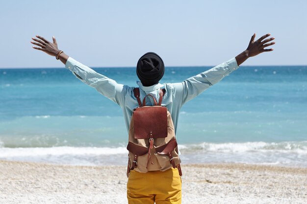 Leggi la vista del felice maschio afroamericano spensierato in piedi sulla spiaggia di fronte al mare azzurro, allargando le braccia, sentendo la libertà e la connessione con la straordinaria natura che lo circonda