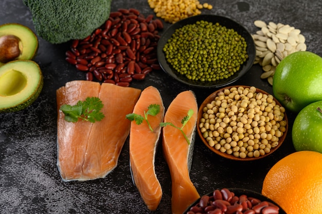 Legumi, broccoli, frutta e salmone posti su un pavimento di cemento nero.