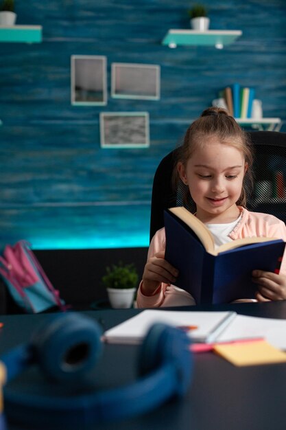 Lettore di Schoolkid che sorride mentre legge un libro educativo che studia per l'esame di letteratura scolastica seduto alla scrivania in soggiorno. Piccolo bambino che lavora ai compiti accademici durante la scuola a casa