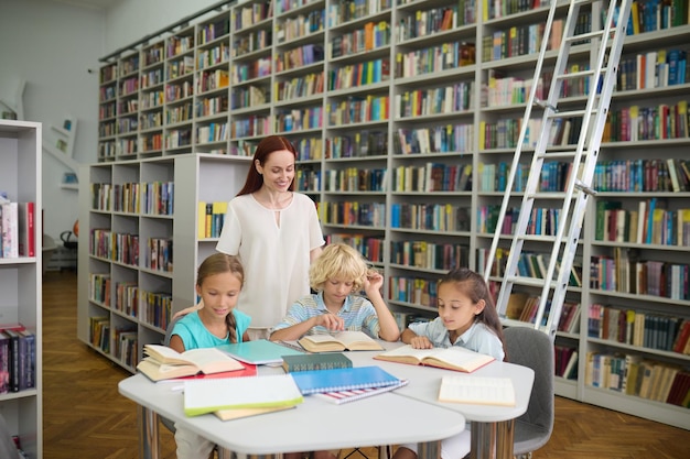 Lettura. Amichevole giovane donna in abiti leggeri in piedi a guardare i bambini della scuola che leggono libri a tavola in biblioteca