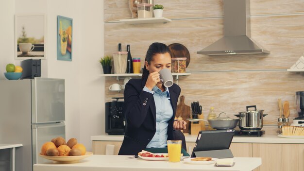 Libero professionista che beve caffè al mattino sul tavolo durante la colazione utilizzando il computer tablet. Donna d'affari che legge le ultime notizie online prima di andare al lavoro, utilizzando la tecnologia moderna in cucina