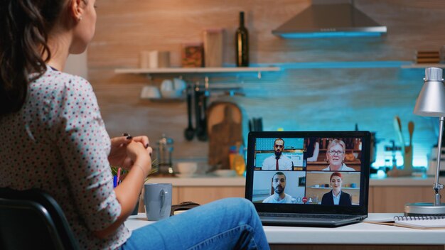 Libero professionista che lavora in remoto discutendo con i partner online utilizzando il laptop seduto in cucina di notte. Utilizzando la moderna tecnologia di rete wireless parlando in una riunione virtuale a mezzanotte facendo gli straordinari