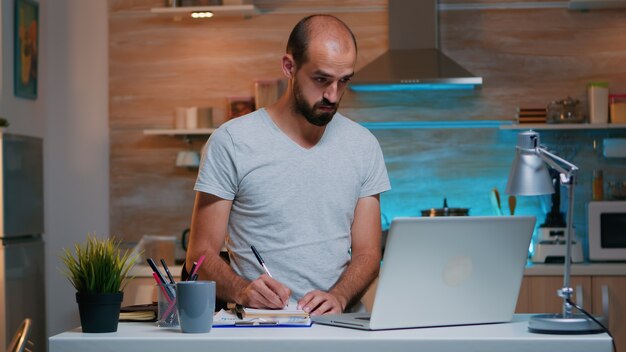 Libero professionista che scrive note nel taccuino mentre studia e utilizza la tecnologia moderna lavorando a casa per gli straordinari. Impiegato impegnato e concentrato che utilizza la moderna tecnologia di lettura wireless della rete digitando, cercando