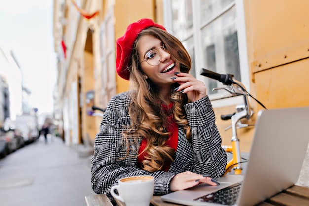 Libero professionista femminile allegro in vetri che posano nella caffetteria all'aperto con computer e bicicletta. Ritratto di donna riccia sorridente in berretto rosso che beve caffè per strada e lavora con il computer portatile.